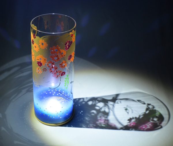 Shown under spot light at upper left, lamp displays brilliant projected color and shadow patterns. Highlighted are displays of patterns through the glass cylinder.
