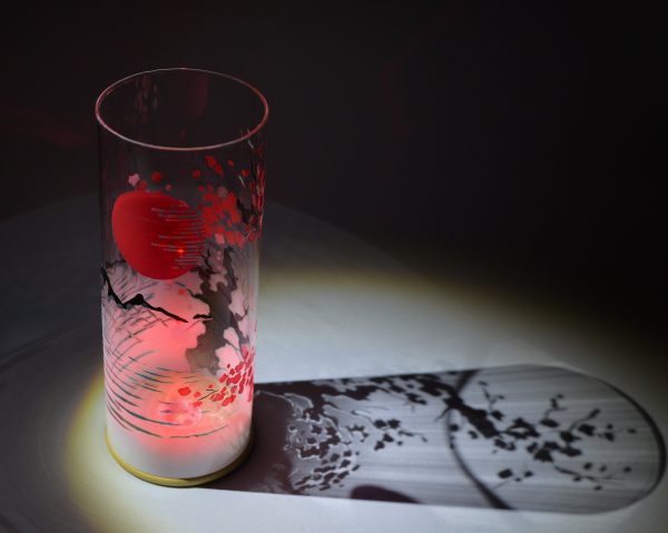 Shown under spot light at upper left, lamp displays brilliant projected shadow patterns. Highlighted are displays of patterns through the glass cylinder. LED set to color red.