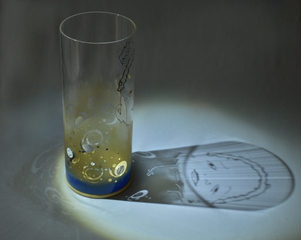 Shown under spot light at upper left, lamp displays brilliant projected shadow pattern of portrait of Jaxson. Highlighted are displays of patterns through the glass cylinder.