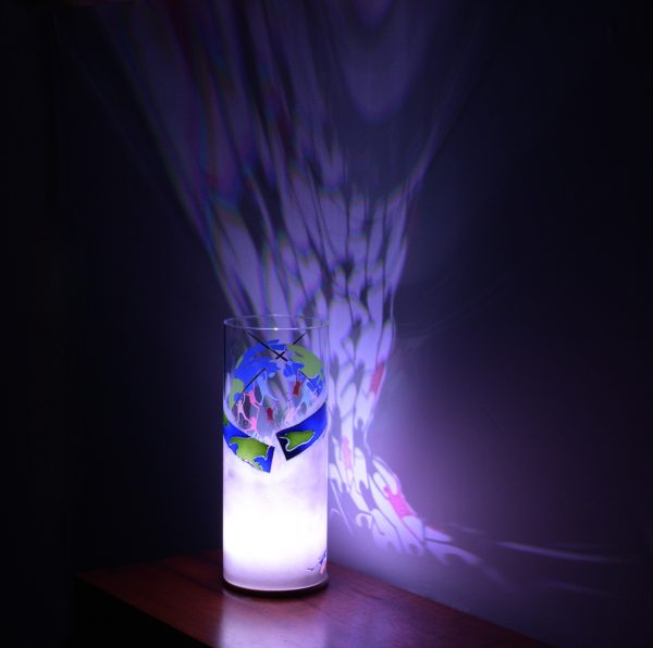 Shown on a display shelf with LED on. Unique art glass lamp displays brilliant patterns of light and shadow onto backdrop.